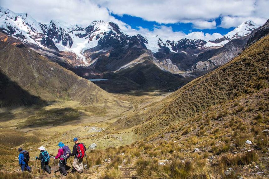 A group of hikers with large backpacks walk along a rugged trail in a mountainous landscape with snow-capped peaks in the background under a partly cloudy sky, embarking on a wild adventure to celebrate someone's 60th birthday in the stunning terrain of Peru.