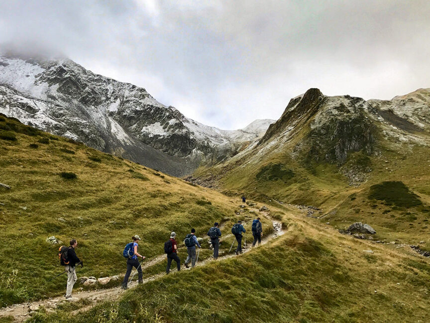 A group of hikers, with their packing lists checked, walks along a mountain trail in the Alps, where grassy slopes and snow-dusted peaks stand majestically beneath a cloudy sky. This adventure unfolds against nature's stunning backdrop.