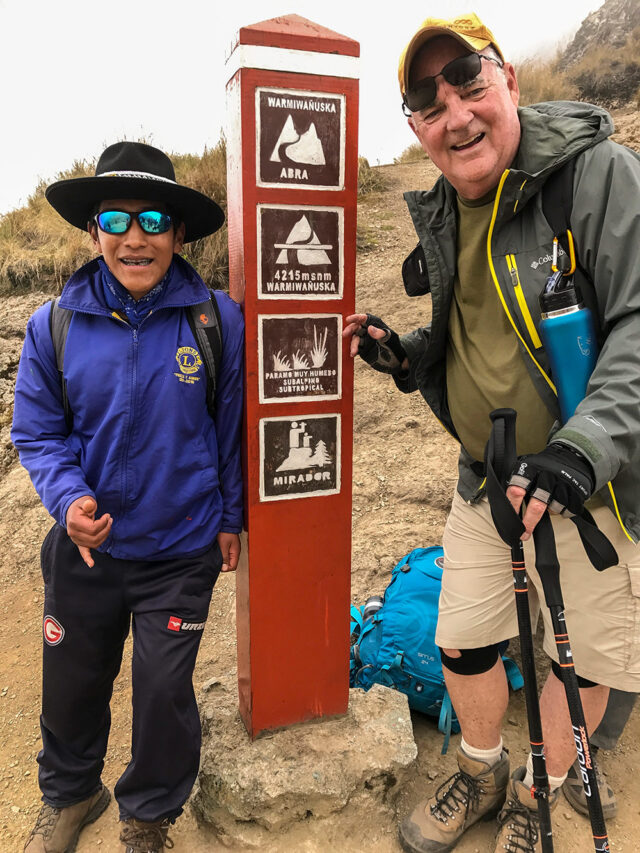Two hikers stand next to a red trail sign indicating "Warmiwañusqa Abra" at 4,215 meters on the Inca Trail. One person is wearing a hat and sunglasses, while the other is wearing shorts and holding trekking poles. Their journey will eventually lead them to the breathtaking site of Machu Picchu.