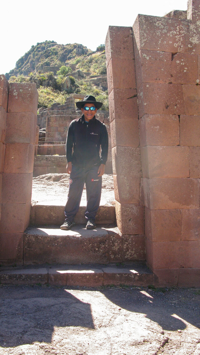 A person wearing sunglasses, a hat, and dark clothing stands between ancient stone structures on the Inca Trail with the mountainous terrain of Machu Picchu in the background.