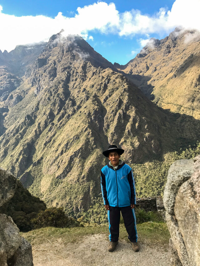         A person wearing a blue tracksuit and black hat stands in front of towering mountains with a cloudy sky above, possibly preparing for hiking the renowned Inca Trail to Machu Picchu.