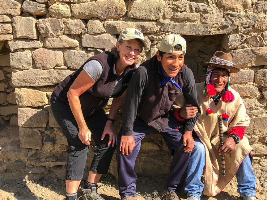 Three people smiling in front of a stone wall. One person plays a wooden flute, wearing a hat adorned with colorful pompoms—a perfect snapshot from their hike along the Inca Trail towards Machu Picchu.
