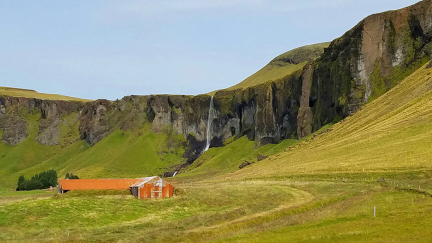 A rusted metal barn sits in a grassy field with a waterfall cascading down a cliff in the background under a blue sky, perfect for hikes exploring Iceland’s rugged landscapes.
