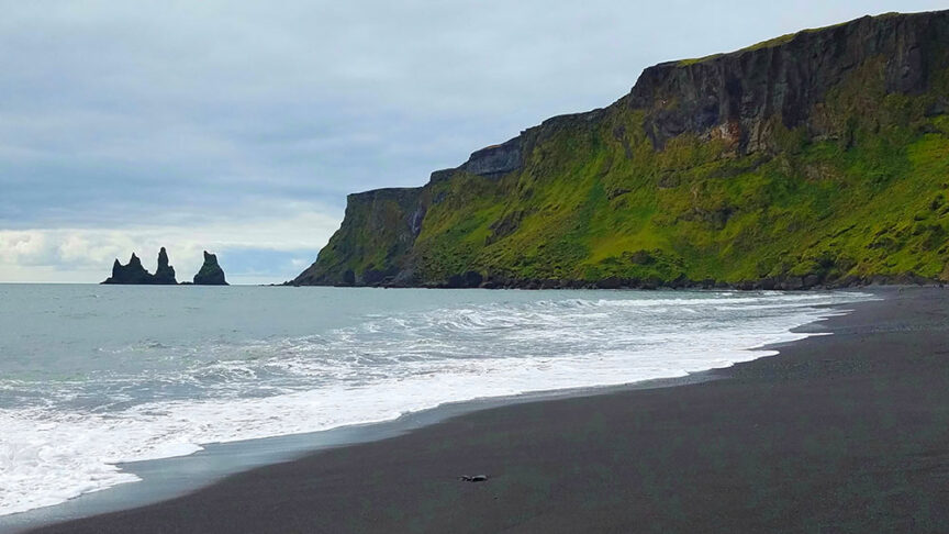 Black sand beach with waves washing ashore, adjacent to a green cliff. Offshore, two rock formations rise from the sea under a cloudy sky. This Icelandic paradise offers scenic hikes and the chance to spot the mesmerizing Northern Lights.