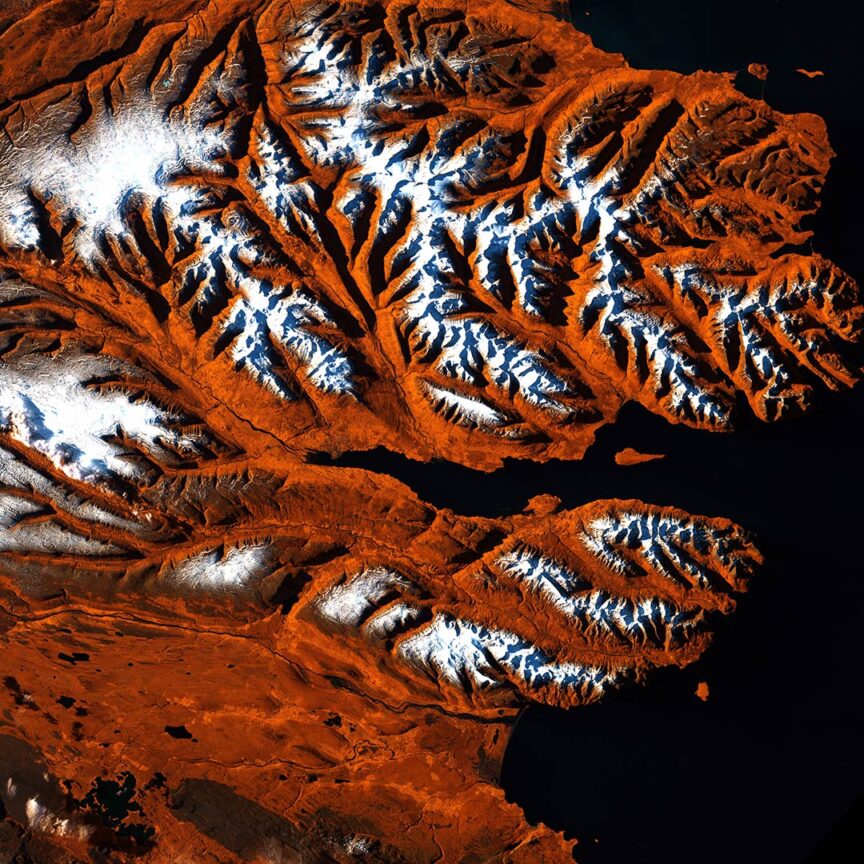 Aerial view of rugged, mountainous terrain with snow-capped peaks contrasted against reddish-brown ground, as captured in stunning satellite images. Water bodies surround and dissect the land, showcasing the raw beauty of Earth from above.