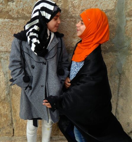 Two young girls in headscarves and coats stand facing each other against a stone wall, one in a gray coat and striped headscarf, the other in an orange headscarf and black shawl, capturing the warmth and beauty of Iran.