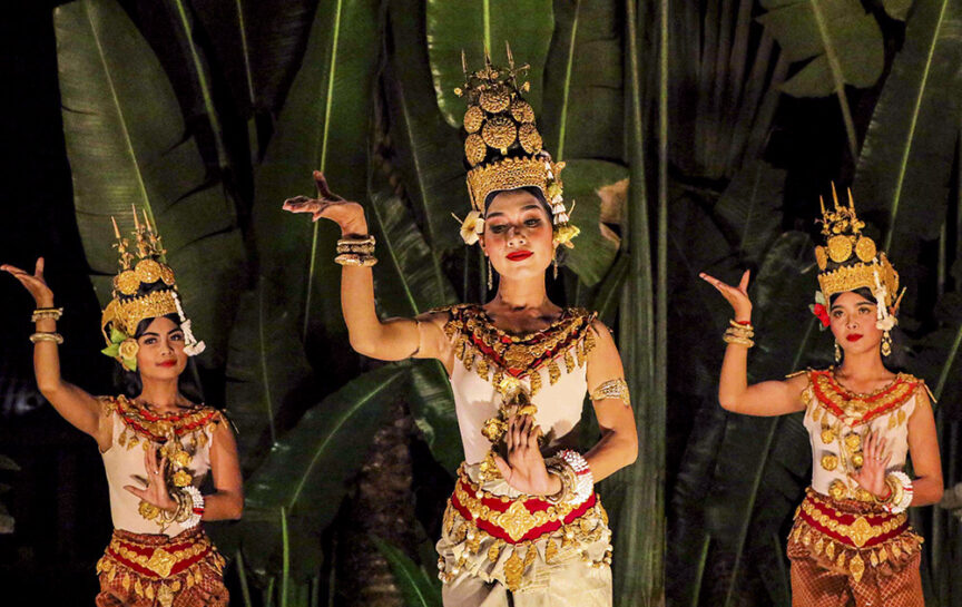 Three women in traditional, ornate costumes perform a cultural dance with intricate hand gestures in front of large green foliage, embodying the charm of "Adventures in Indochine.