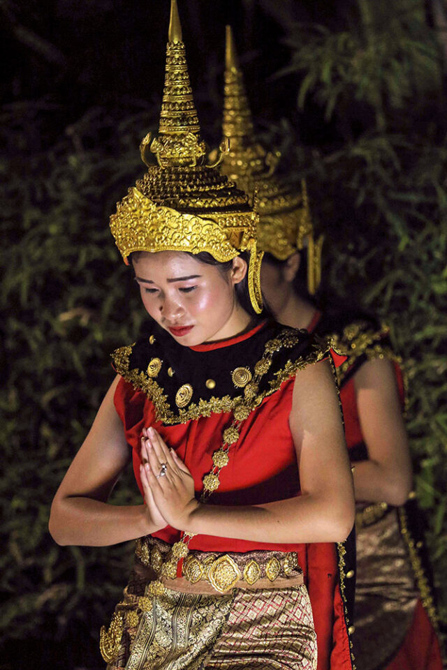 Two people in traditional Thai clothing and gold headdresses, with the person in the foreground performing a dance gesture with hands together. Their graceful movements encapsulate the spirit of 'Adventures in Indochine,' offering a glimpse into the rich cultural tapestry often described as generating 'Five Hundred Smiles.'