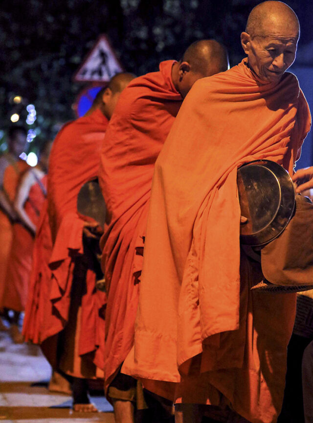 A group of monks dressed in orange robes stand in a line, carrying black alms bowls, embodying the serene spirit of Adventures in Indochine.