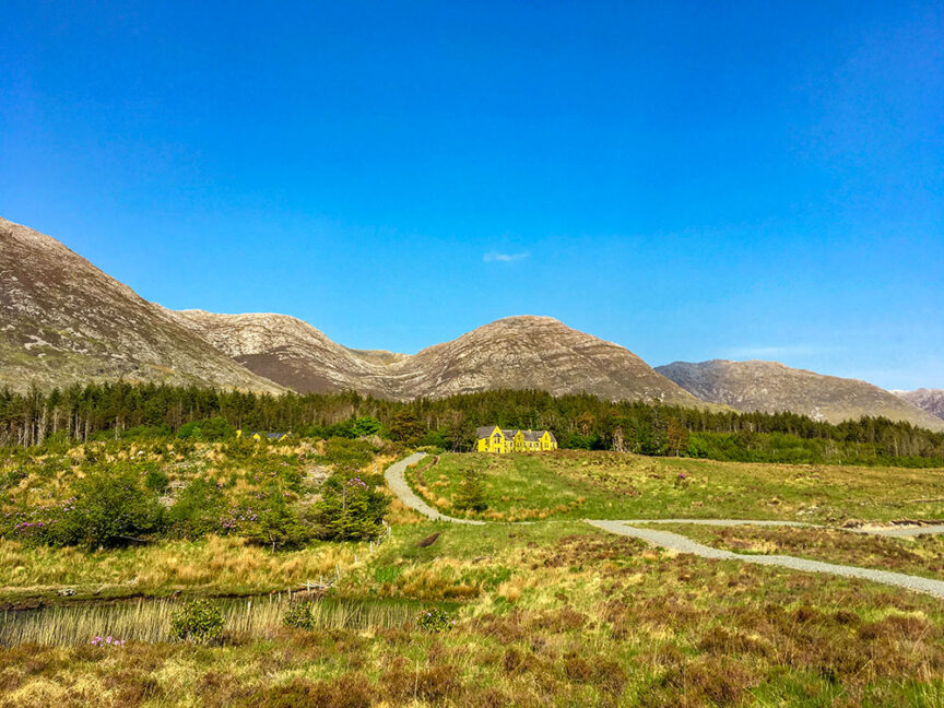 A yellow house is nestled in a green valley with a winding path leading to it, surrounded by grassy plains and trees. Tall, rocky peaks stand majestically in the background under a clear blue sky, reminiscent of Ireland's West Coast.