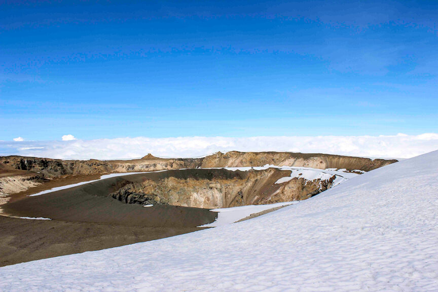 A snow-covered landscape with a large crater in the center under a clear blue sky reminds me of my mother’s stories about climbing Kili. The horizon shows a beautiful layer of clouds, just as she described from her adventures.