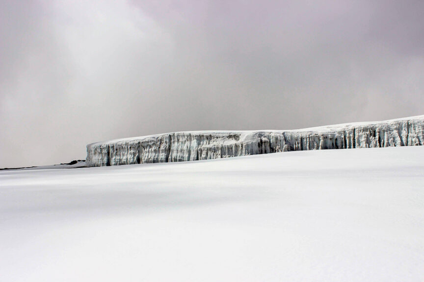 The image shows a vast, snowy landscape with a large ice cliff extending horizontally under a cloudy sky, reminiscent of stories Grandmother used to tell about her adventurous days climbing Kili.