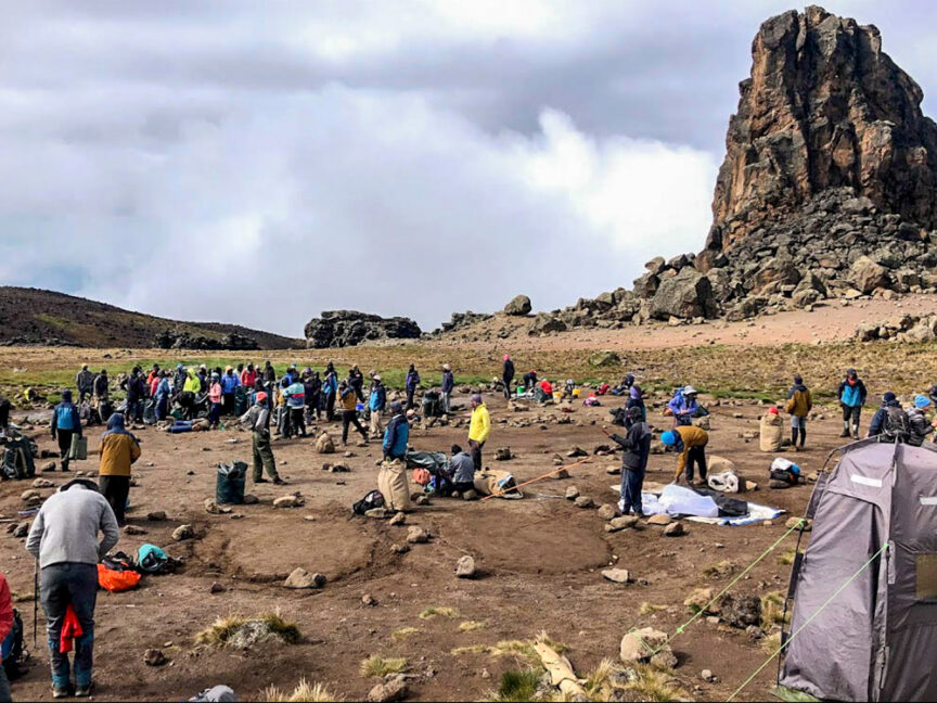 A large group of people, led by a determined grandmother, set up camp in a rocky, open area with tents and supplies spread out on the ground. A prominent rock formation stands in the background under a cloudy sky, reminiscent of her epic stories about climbing Kili.