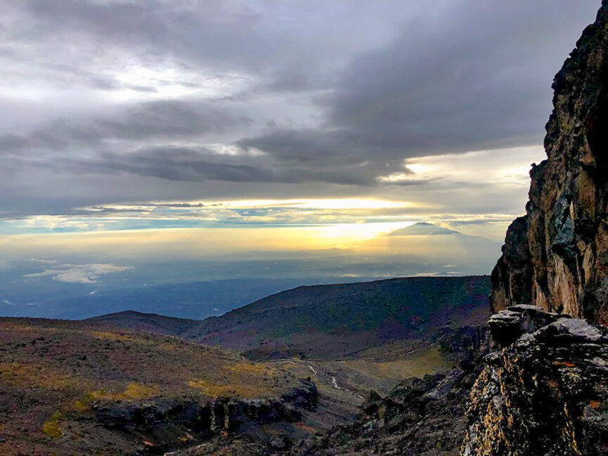 Sunrise view over a mountain range with a cloudy sky and rugged terrain, reminiscent of tales from my grandmother's adventures. Rocks and cliffs are visible in the foreground, inviting explorers as if ready for Climbing Kili.