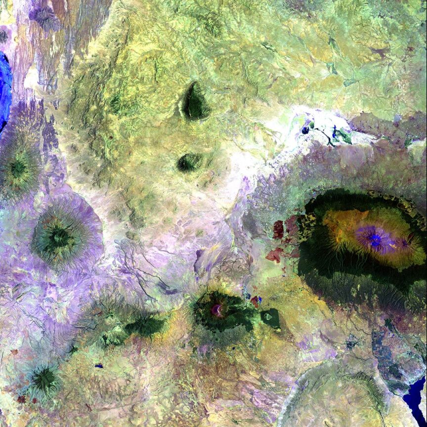 A stunning satellite view shows a diverse and colorful terrain featuring various shades of green, brown, and blue, with prominent irregular shapes and patterns resembling natural landforms or geological features.