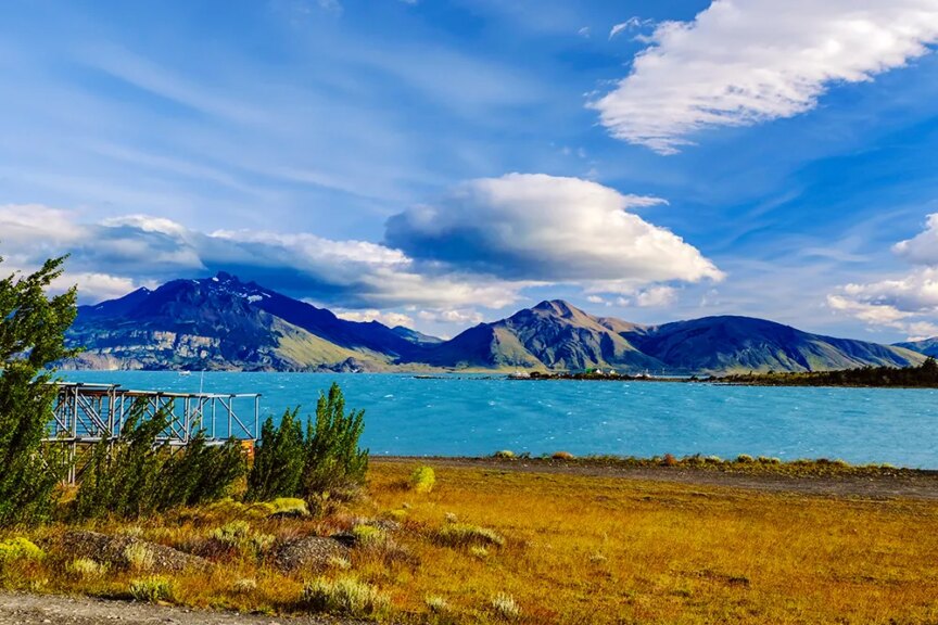A serene lake with turquoise water is bordered by yellowish-brown grass, evoking impressions of untouched beauty, with mountains in the background under a partly cloudy Patagonian sky.