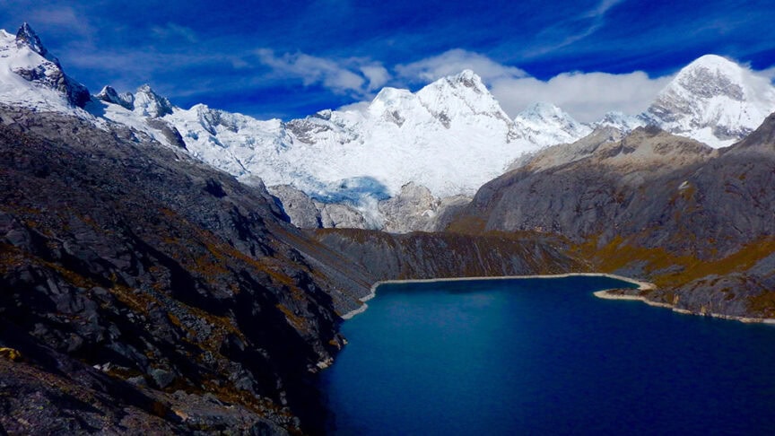 Panoramic view of a deep blue lake surrounded by rugged, snow-covered mountains under a bright blue sky with scattered clouds, perfect for celebrating a 60th birthday in the wilds of Peru.