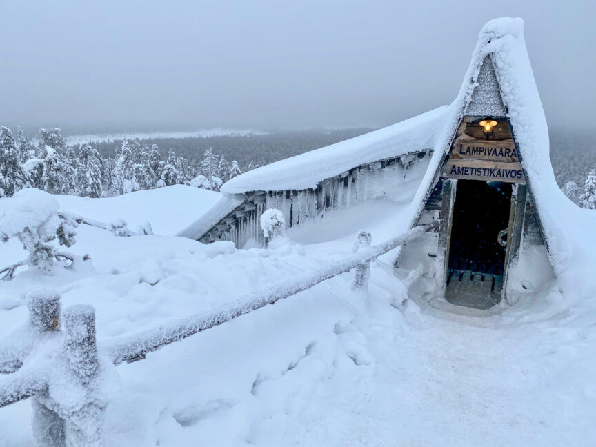 A snow-covered structure with a steep roof and a sign reads "Lampivaara Amethyst Mine" amid a snowy, foggy landscape. A narrow pathway leads to the entrance, inviting you on an adventure in the heart of Northern Finland.