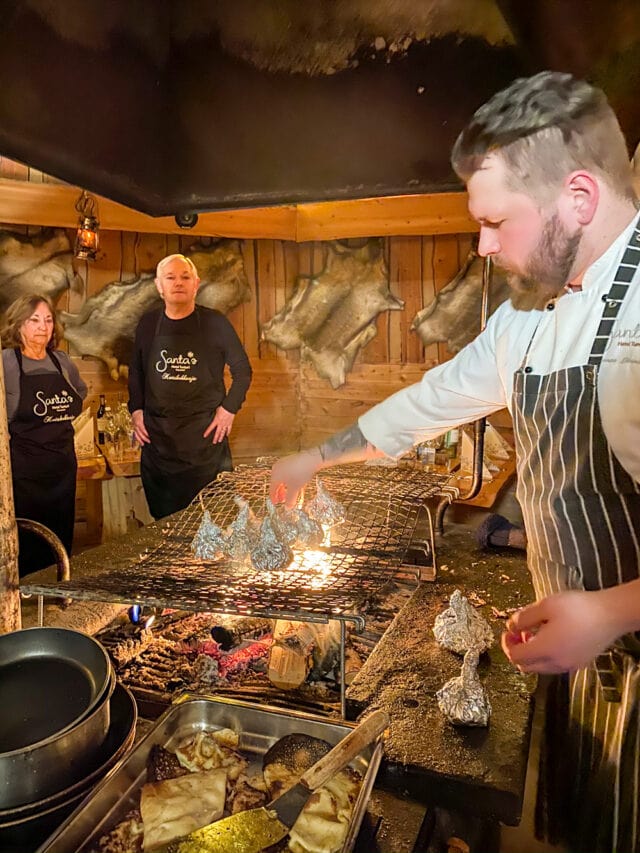 In a rustic setting, a chef enthralls with culinary adventures over an open flame, while two onlookers observe. Food items wrapped in foil sizzle on the grill, evoking the spirit of Northern Finland.
