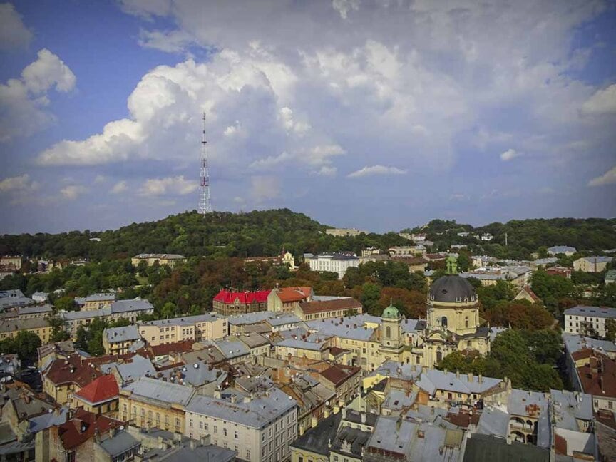 Aerial view of a city with a large cathedral building, historic roofs, and a hill with a broadcasting tower in the background under a partly cloudy sky, evoking memories from my search of my roots during the Great Carpathian Traverse.