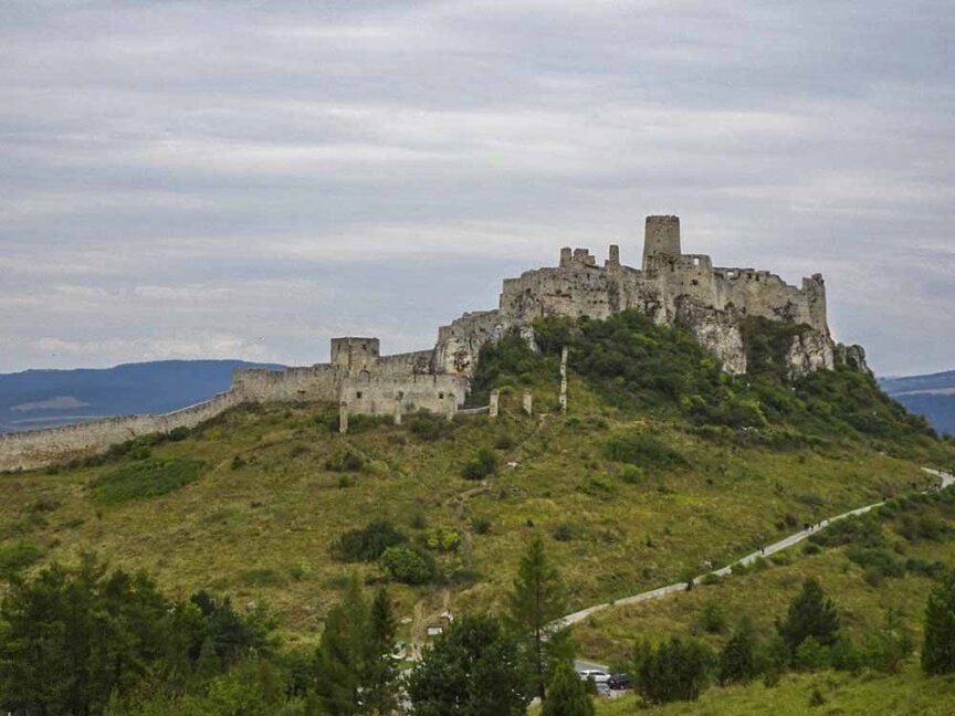 A medieval castle sits atop a grassy hill, surrounded by a stone wall. A winding path leads to the entrance amid a backdrop of distant mountains and a cloudy sky, much like the scenic vistas one might encounter on the Great Carpathian Traverse in search of my roots.