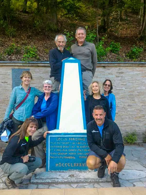 A group of eight adults pose around a tall, blue and white obelisk with inscriptions, their faces glowing with the excitement of their Carpathian Traverse adventure. In the background, lush trees whisper tales of roots that intertwine history and nature.