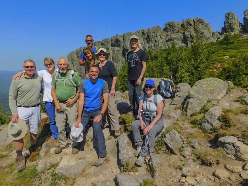 A group of nine hikers poses on a rocky trail during their Carpathian Traverse, framed by a scenic mountain backdrop under a clear blue sky.