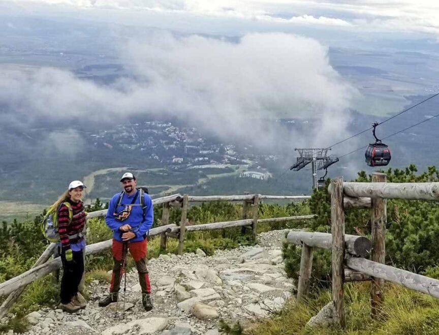 Two hikers with backpacks and trekking poles stand on a rocky path next to a wooden fence, embarking on the Great Carpathian Traverse. A cable car and a cloudy landscape with buildings and fields are visible in the background.