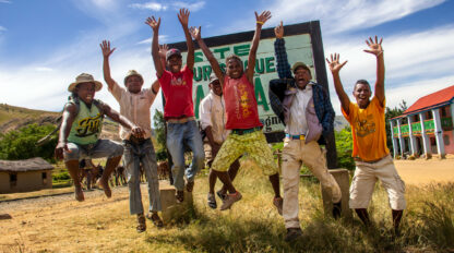 A group of Wilderness Travelers jumps joyfully in the air in front of a sign in an outdoor setting with buildings and clear skies in the background.