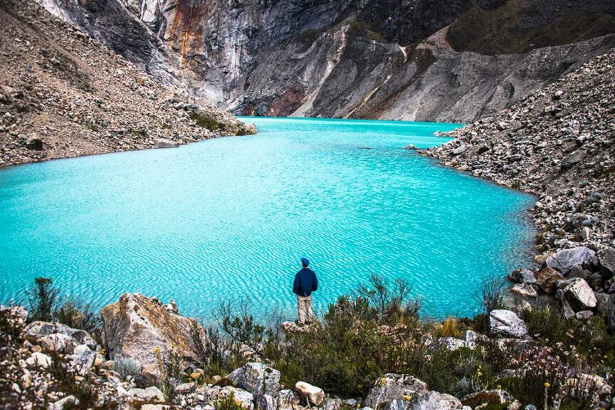 A person in a blue jacket stands at the edge of a vibrant turquoise lake in the wilds of Peru, surrounded by rocky mountains and sparse vegetation, celebrating their 60th birthday.