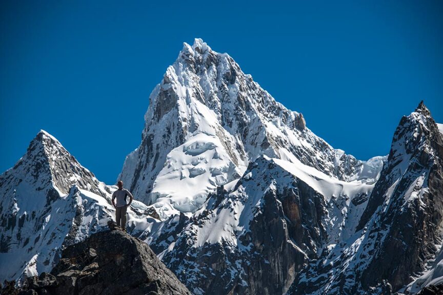 A person stands on a rocky outcrop, gazing at the snow-covered mountain peaks of Peru under a clear blue sky, perhaps in celebration of their 60th.