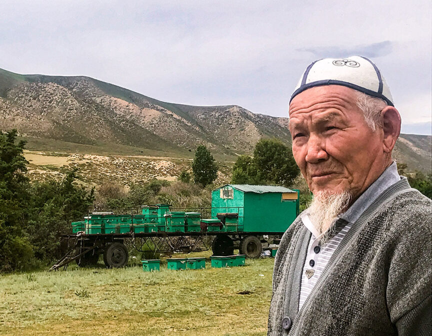 An elderly man stands in a rural landscape with green hills in the background and a green beekeeping trailer and boxes behind him. He wears a traditional hat and a grey cardigan, evoking the timeless essence of cultural journeys along the Silk Road.