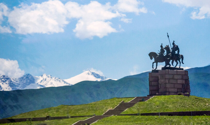 Two silhouetted horsemen statues on a raised platform against a backdrop of green mountains and snowy peaks under a bright blue sky with clouds, capturing the essence of a historic Silk Road cultural journey.