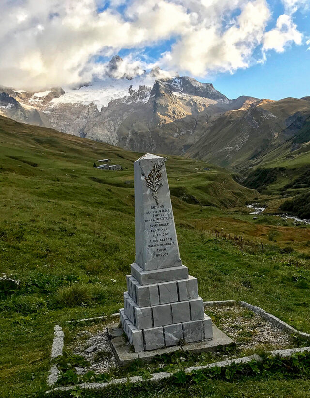 A stone monument with inscriptions stands in a grassy landscape, set against the backdrop of snow-capped peaks partially veiled by clouds, reminiscent of views along the Tour du Mont Blanc hiking trail.