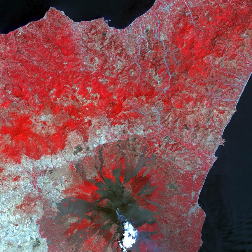 Stunning satellite image showing a large volcanic mountain with red-tinted areas indicating vegetation or geological features. The volcano has a distinct crater, while surrounding areas display varied terrain and waterways, showcasing Earth's diverse landscape.