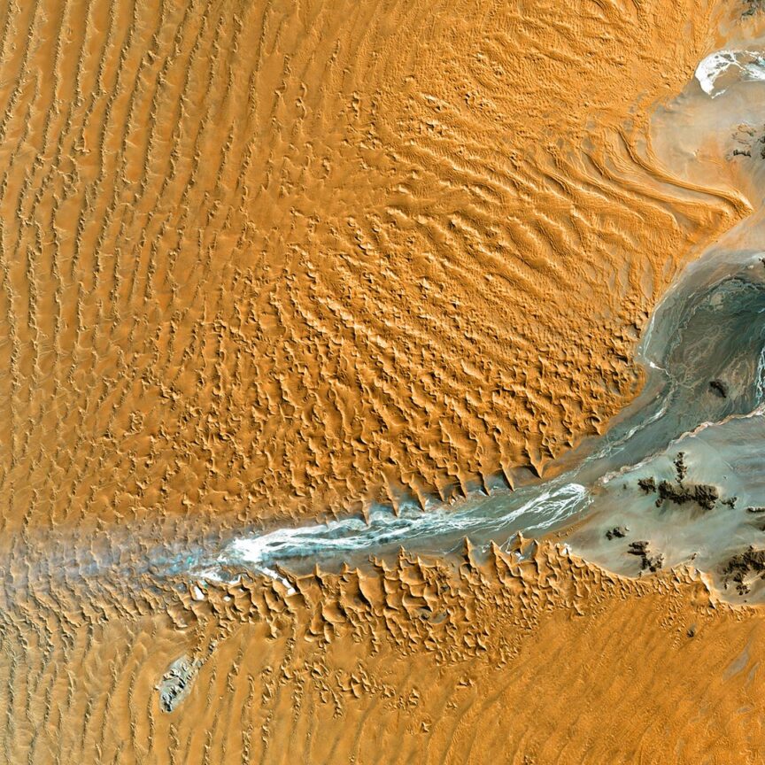 Aerial view of a desert landscape with rippled sand dunes and a narrow stream of water flowing through the middle, partially surrounded by wet patches. This stunning scene could easily be mistaken for one of Earth's satellite images.