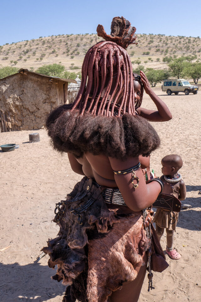 A woman in traditional attire and distinctive hair stands outdoors in a rural area of Namibia, accompanied by a child. A thatched-roof structure and a vehicle are visible in the background, blending seamlessly into the Namibian landscapes teeming with wildlife.