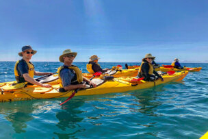 A group of people wearing hats and life jackets paddle yellow kayaks on calm, blue water under a clear New Zealand sky, with majestic mountains in the distance.
