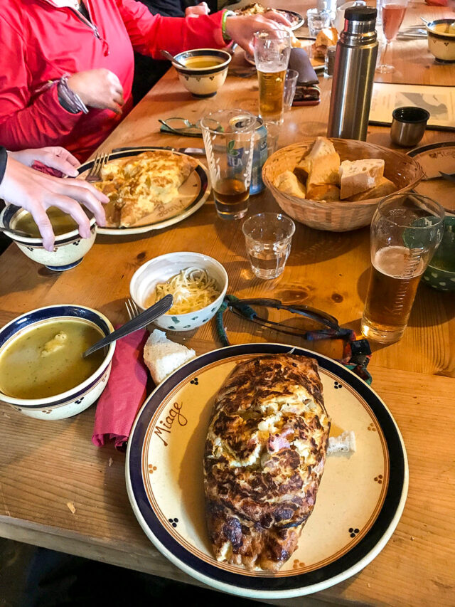A table with three plates of food, assorted glasses of drinks, a basket of bread, soup bowls, and utensils. People are reaching for food after hiking the Tour du Mont Blanc. One plate has a grilled item with melted cheese.