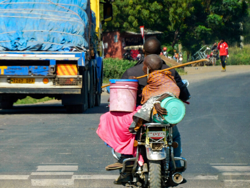 Two people ride a motorcycle on a road, one carrying buckets; a truck is ahead on the left. Trees and another person are visible in the background, evoking a scene from a Serengeti Safari.