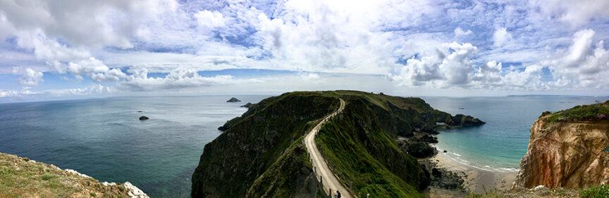 Panoramic view of Britain's Land with a rugged coastal landscape, a narrow road winding through lush green hills under a cloudy sky, perfect for hiking and feeling lost in time, surrounded by the sea.