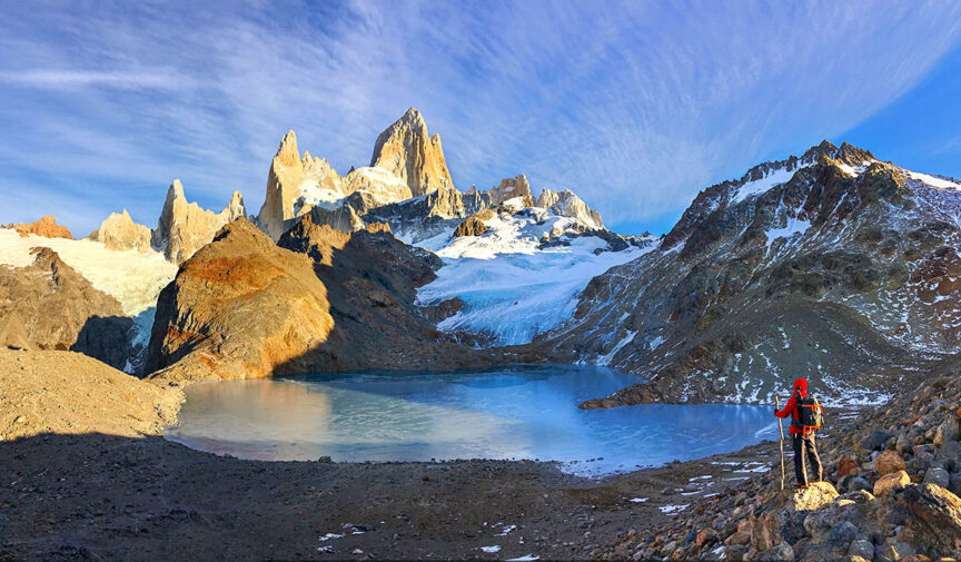 A person in hiking gear stands at the edge of a glacial lake with rugged snow-capped mountains in the background under a clear blue sky, embodying the serenity that makes trips to book so worth planning in advance.