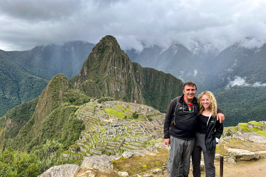 Two people stand smiling in front of the ancient Inca city of Machu Picchu, nestled in the lush green mountains and cloudy skies of Peru's Sacred Valley.