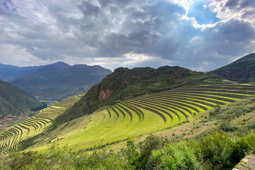 Terraced fields on a hillside under a partly cloudy sky with distant mountains and valleys in the background, reminiscent of the stunning landscapes found in Peru's Sacred Valley.