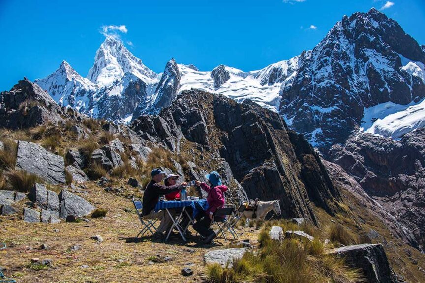 Three people seated at a small table set with food and drinks, celebrating in Peru amidst the wilds of Peru, enjoying a meal outdoors with towering snow-covered mountains in the background.