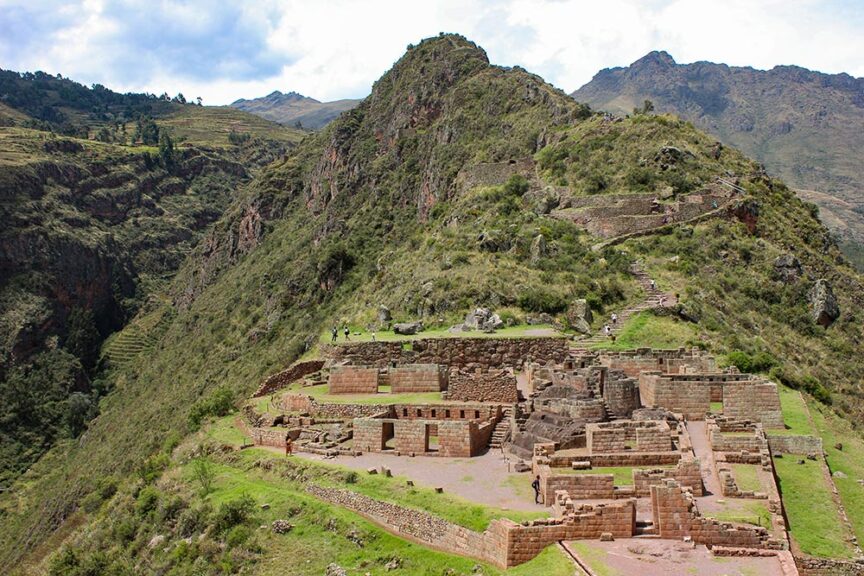 Following in the footsteps of the Incas, ancient stone ruins are nestled in a mountainous landscape with terraced hillsides and a cloudy sky above.