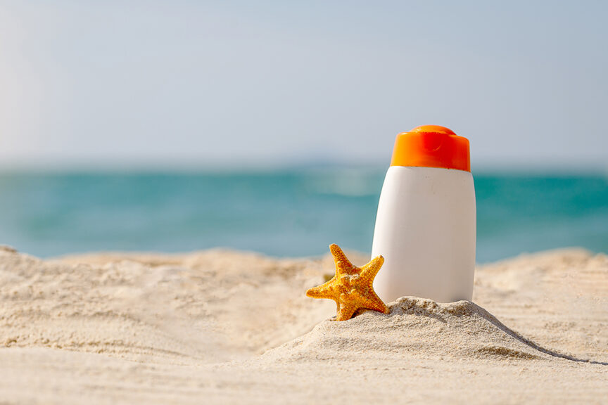 A bottle of sunscreen and a starfish on a sandy beach with the ocean in the background under a clear sky—perfect items to remember for your next snorkeling trip.