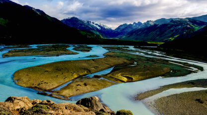 A winding river with multiple channels and islands flows through a mountainous landscape in Patagonia, leaving lasting impressions under a partly cloudy sky.