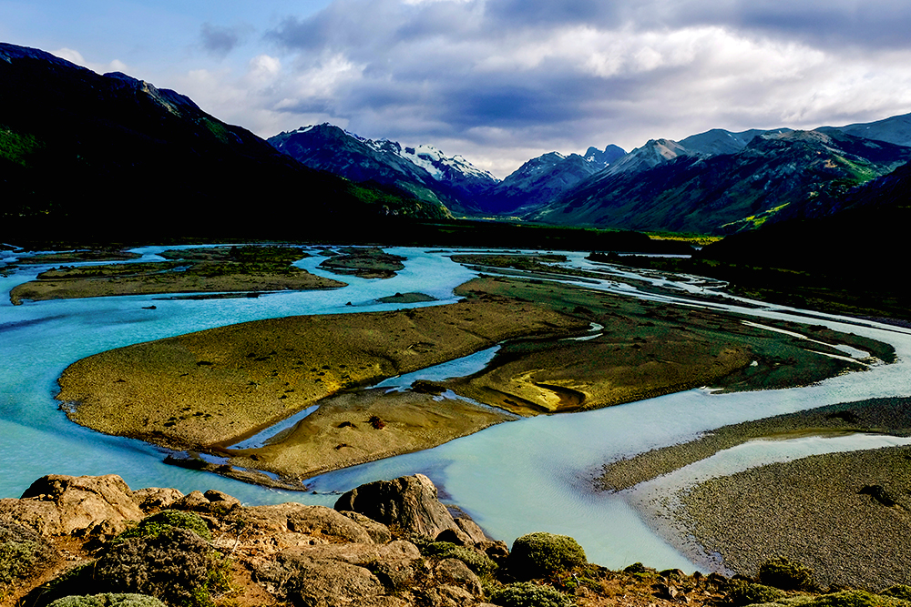 A winding river with multiple channels and islands flows through a mountainous landscape in Patagonia, leaving lasting impressions under a partly cloudy sky.