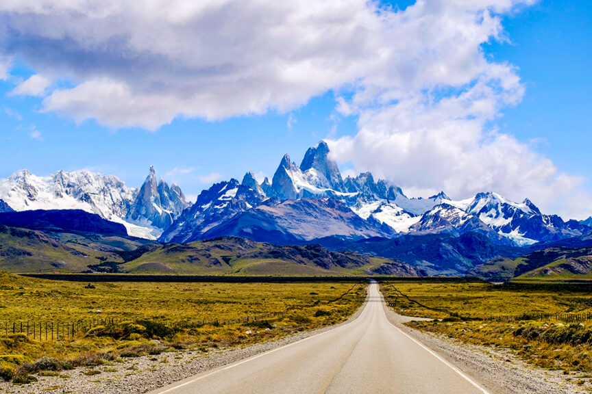 A straight road leads towards the snowy mountains of Patagonia under a partly cloudy sky, surrounded by grassy plains, leaving lasting impressions of nature's grandeur.